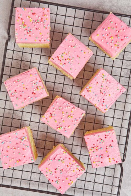 Colorful frosted sugar cookie bars with pink icing and rainbow sprinkles on a wire cooling rack.