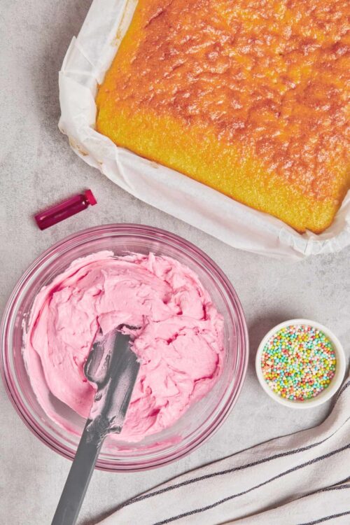 Fluffy pink frosting in a glass bowl next to a baked cake with colorful sprinkles on a textured surface.