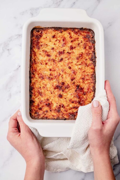 Savory baked casserole in white dish with hands holding edges on marble surface.