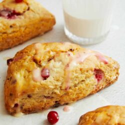 Freshly baked cranberry orange scones drizzled with glaze with a glass of milk on a white surface.