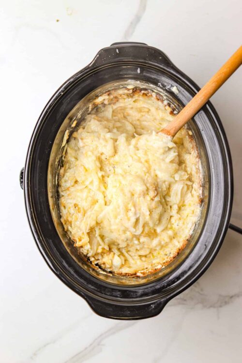 Creamy mashed potatoes in a slow cooker with a wooden spoon in a black crockpot.