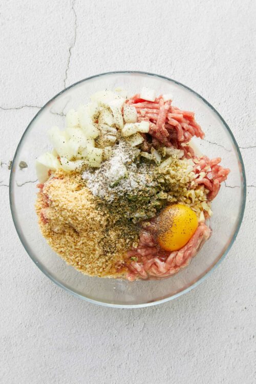 Minced meat mixture with eggs, breadcrumbs, chopped onions, seasonings, and ground meat in a glass bowl.