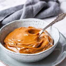 Creamy caramel sauce in a rustic ceramic bowl on a matching plate, with a silver spoon and a soft gray cloth in the background.