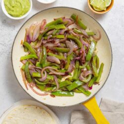 Sauteed onions and green peppers in a yellow skillet with guacamole, sour cream, and lime slices on white background.
