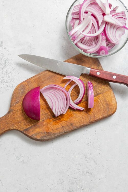 Sliced red onion on wooden cutting board with knife and bowl of onion rings, fresh and ready for cooking or salad.