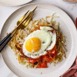 Golden fried egg on hash browns with sautéed tomatoes, served on a textured white plate.