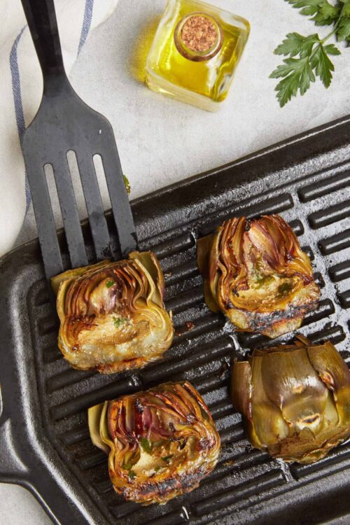 Roasted artichokes in cast iron skillet with olive oil and herbs on a kitchen countertop.