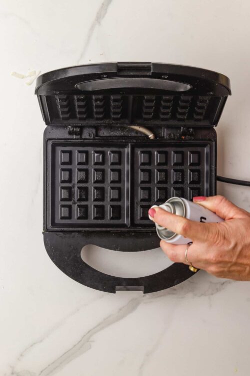 Golden-brown waffle being sprayed with non-stick cooking spray in the waffle maker.