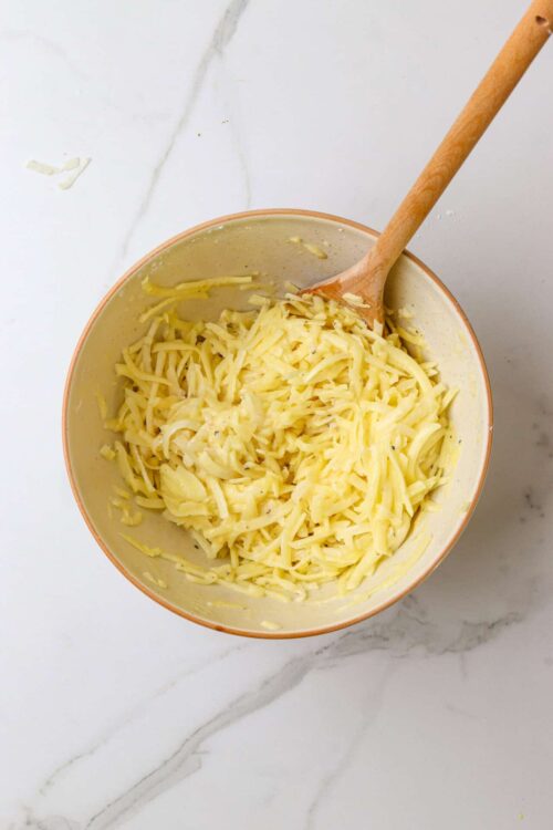 Creamy shredded cheese mixture in a bowl with a wooden spoon on a white marble surface.