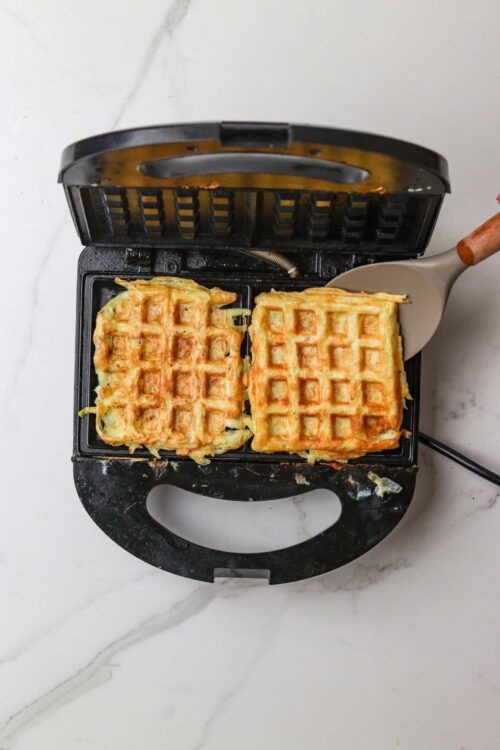 Crispy waffles being made in a waffle maker with batter on top, on a white marble surface.