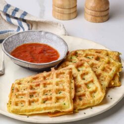Crispy waffles with chives and a side of tomato sauce, served on a white plate, food photography.