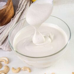 Creamy homemade white sauce in a glass bowl, with lemon and cashews on a white cloth.