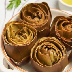 Roasted artichokes with a golden brown exterior served on a white platter with lemon herb dipping sauce.