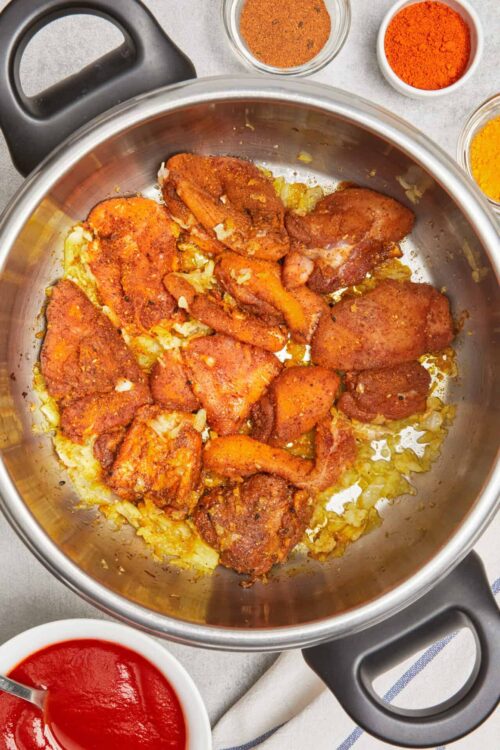 Crispy seasoned chicken thighs cooking in a stainless steel skillet with spices and onions.