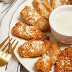 Crispy fried chicken tenders with seasoned coating served with creamy dipping sauce on a white plate.