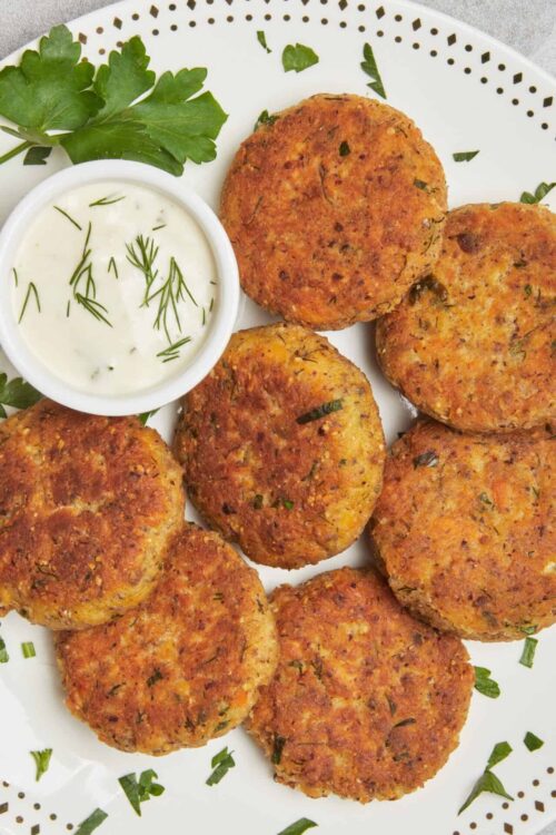 Crispy salmon cakes with dill yogurt sauce and fresh herbs on white plate.