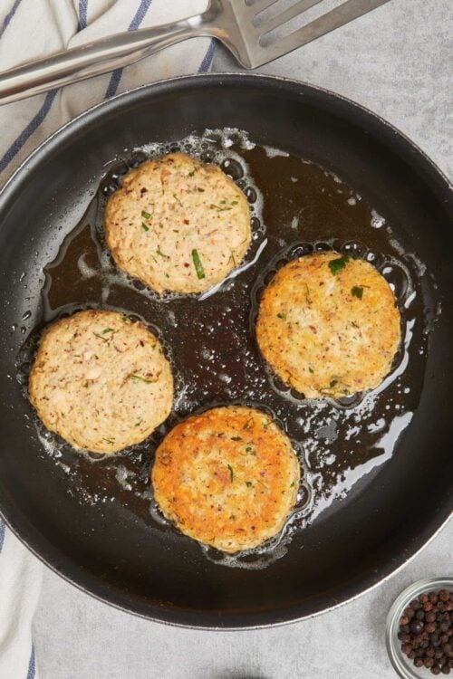 Golden crab cakes sizzling in a skillet with herbs and spices.