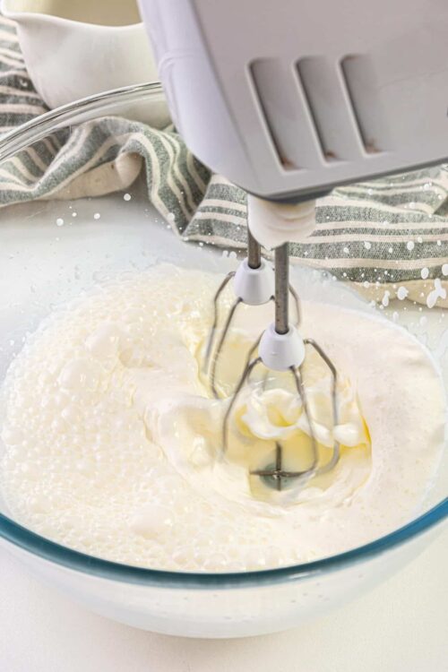 Fluffy whipped cream being mixed with an electric hand mixer in a glass bowl.