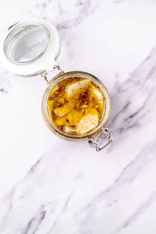 Pickled garlic cloves in a glass jar with brine on a white marble surface.