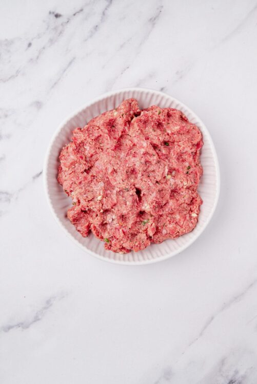 Minced meat mixture for homemade sausage or meatball preparation on a white plate on marble surface.