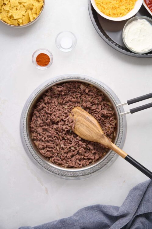 Cooked ground beef in a stainless steel skillet with a wooden spoon, surrounded by bowls of shredded cheese, sour cream, and taco ingredients.