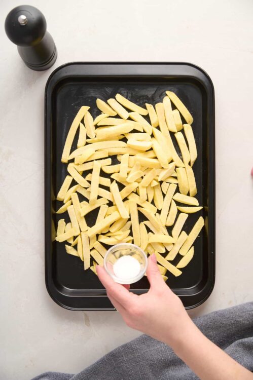 Baked French fries on a baking sheet with salt being sprinkled.