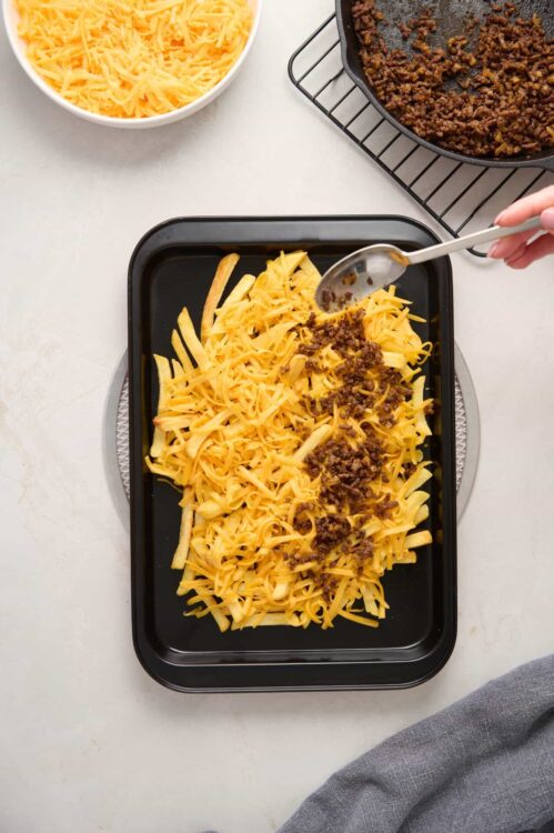 Shredded cheese and cooked ground beef layered in a baking dish for lasagna preparation.