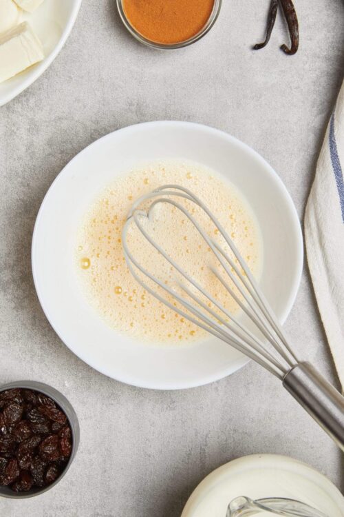 Whisked eggs in a white bowl for baking recipes, with ingredients around on textured surface.