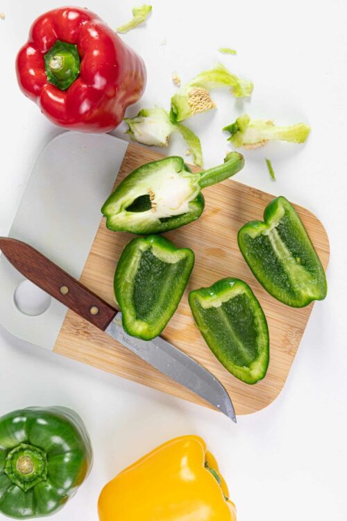 Vibrant green, yellow, and red bell peppers on a wooden cutting board with a knife.