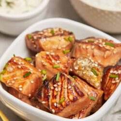Succulent glazed salmon bites topped with sesame seeds and chives in a white dish.