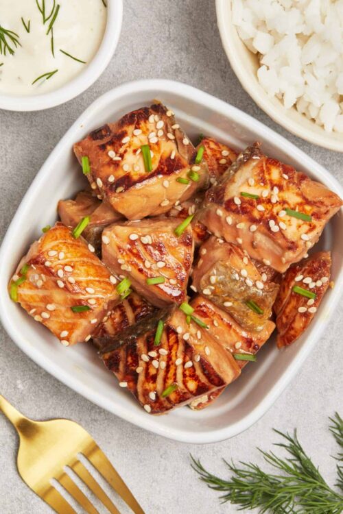 Succulent grilled salmon bites topped with sesame seeds and chopped green onions on a white dish.