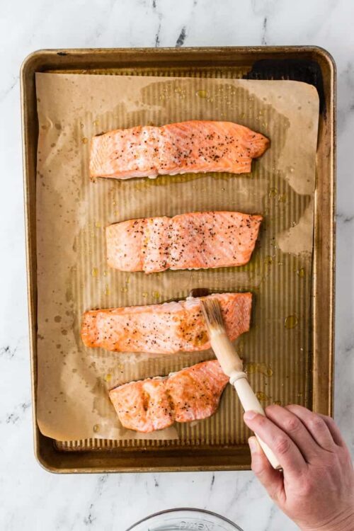 Salted salmon fillets on baking sheet, seasoned with black pepper and drizzled with olive oil.