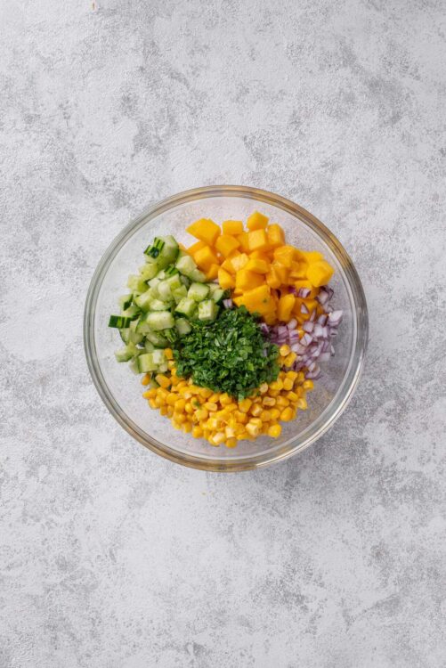 Chopped vegetables in a glass bowl, including cucumber, mango, red onion, chopped cilantro, and corn for salad or wraps.