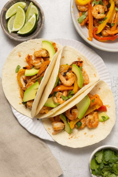 Grilled shrimp tacos with avocado and bell peppers on a white plate, fresh lime wedges on the side.