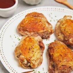 Juicy baked chicken thighs with crispy skin on white plate, with dipping sauces and green garnish.