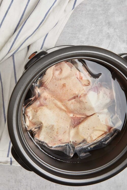 Raw chicken in a slow cooker with a glass lid and plastic wrap, ready to cook.
