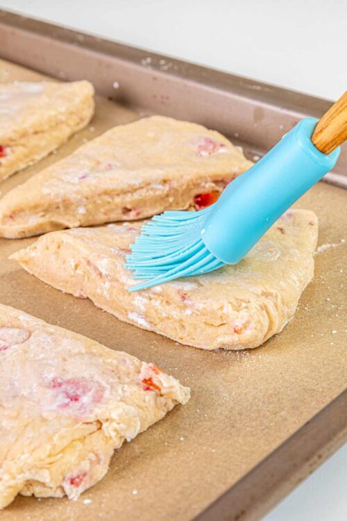 Buttery strawberry scones being brushed with egg wash before baking in an oven.
