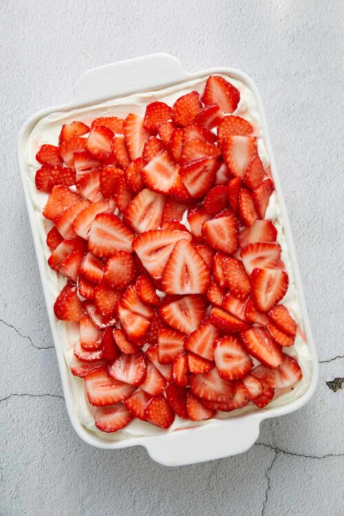 Fresh strawberry shortcake with whipped cream in a white baking dish, topped with sliced strawberries.