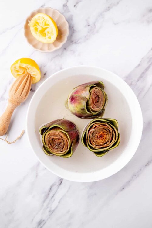 Vegan artichoke hearts on a white plate with lemon wedges and a citrus juicer on marble surface.