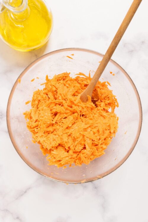 Shredded cheddar cheese in a glass bowl with a wooden spoon, on a marble surface.