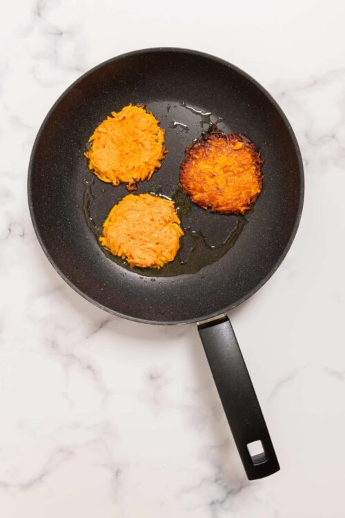 Crispy sweet potato pancakes in a frying pan on marble countertop.