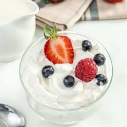 Fresh strawberry and berry-topped whipped cream in a glass bowl on a white surface with additional strawberries and cream in the background.