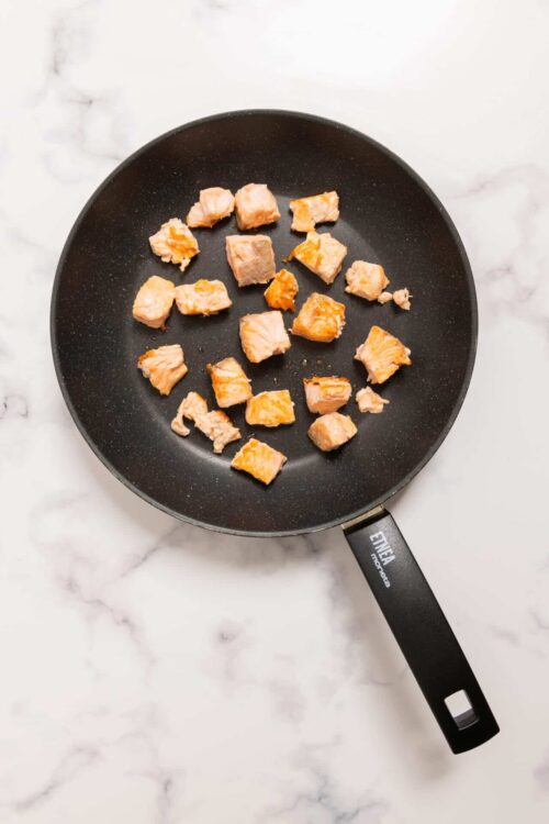 Sautéed salmon pieces in a non-stick frying pan on marble countertop.