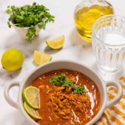 Rich shredded chicken tortilla soup with lime wedges, fresh cilantro, and crunchy white and yellow striped dish towel on marble surface.