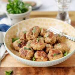 Juicy homemade meatballs in a decorative bowl on a wooden cutting board with fresh herbs.