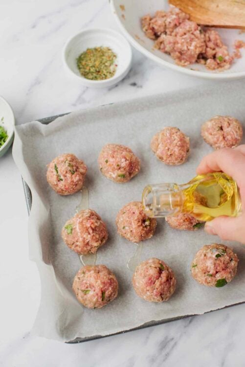Minced meatball mixture forming into balls on baking sheet with olive oil being drizzled, food prep, cooking, baking process.