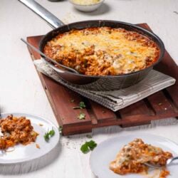 Cheesy baked lasagna in a cast iron skillet with fresh herbs and side dishes on a white rustic table.