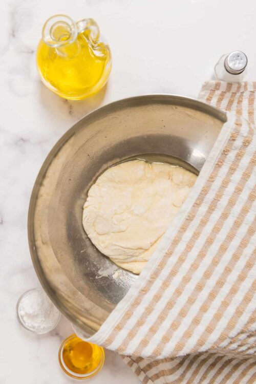 Softened pizza dough resting in a mixing bowl with a striped towel, olive oil, and salt shaker nearby.