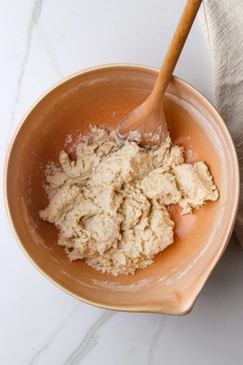 Buttery pie crust dough in a peach-colored bowl with a wooden spoon for mixing.