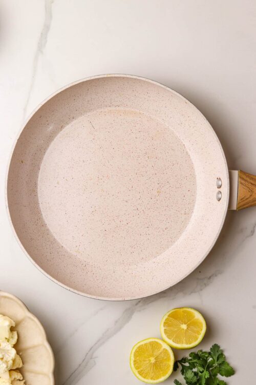 Stoneware frying pan with a speckled beige interior on marble surface, lemon and cilantro garnish nearby.